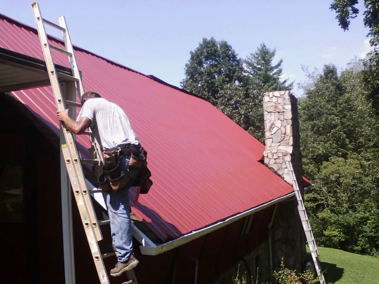 Roofer on a ladder beside a red metal roof and stone chimney — Soffit & Fascia Repair by Roofing Brothers in Louviers